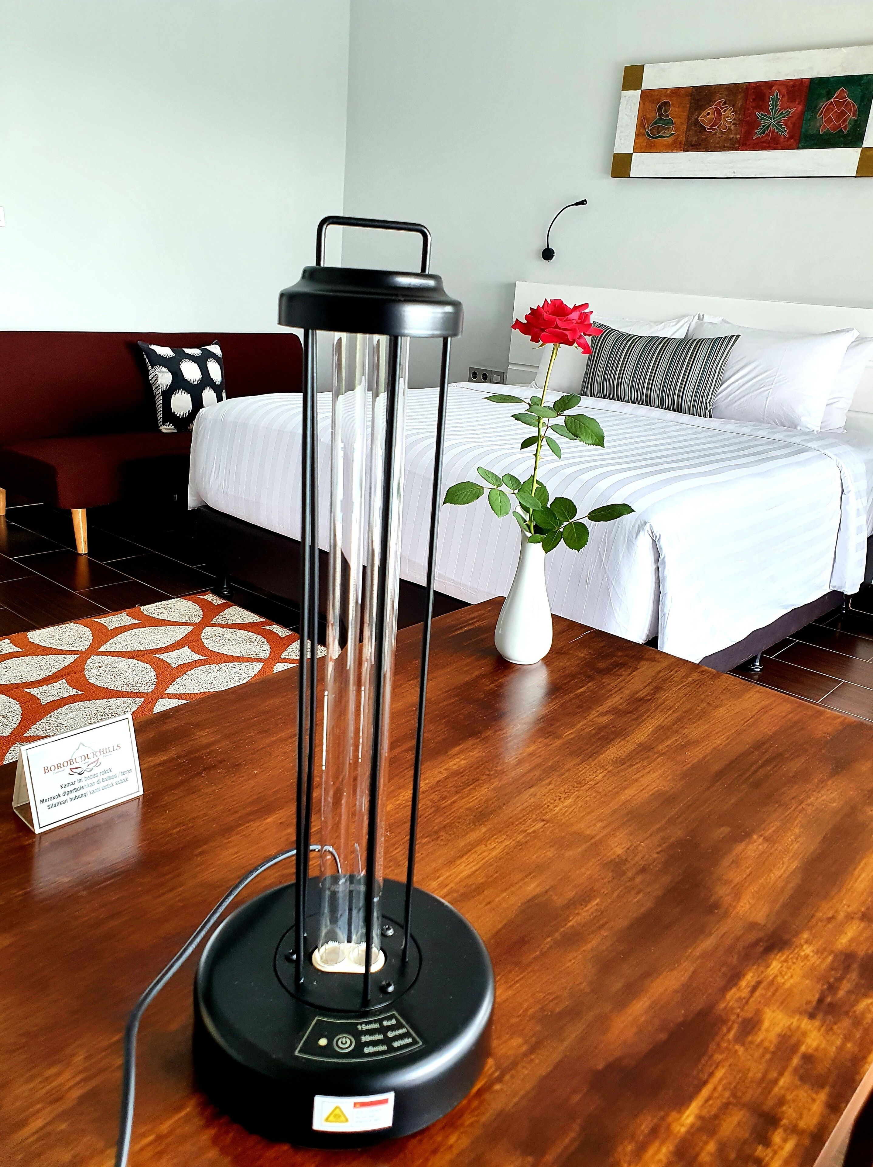 A modern hotel room with a wooden floor, a bed with white bedding, a red sofa, a white vase with a red rose, and a black portable heater.