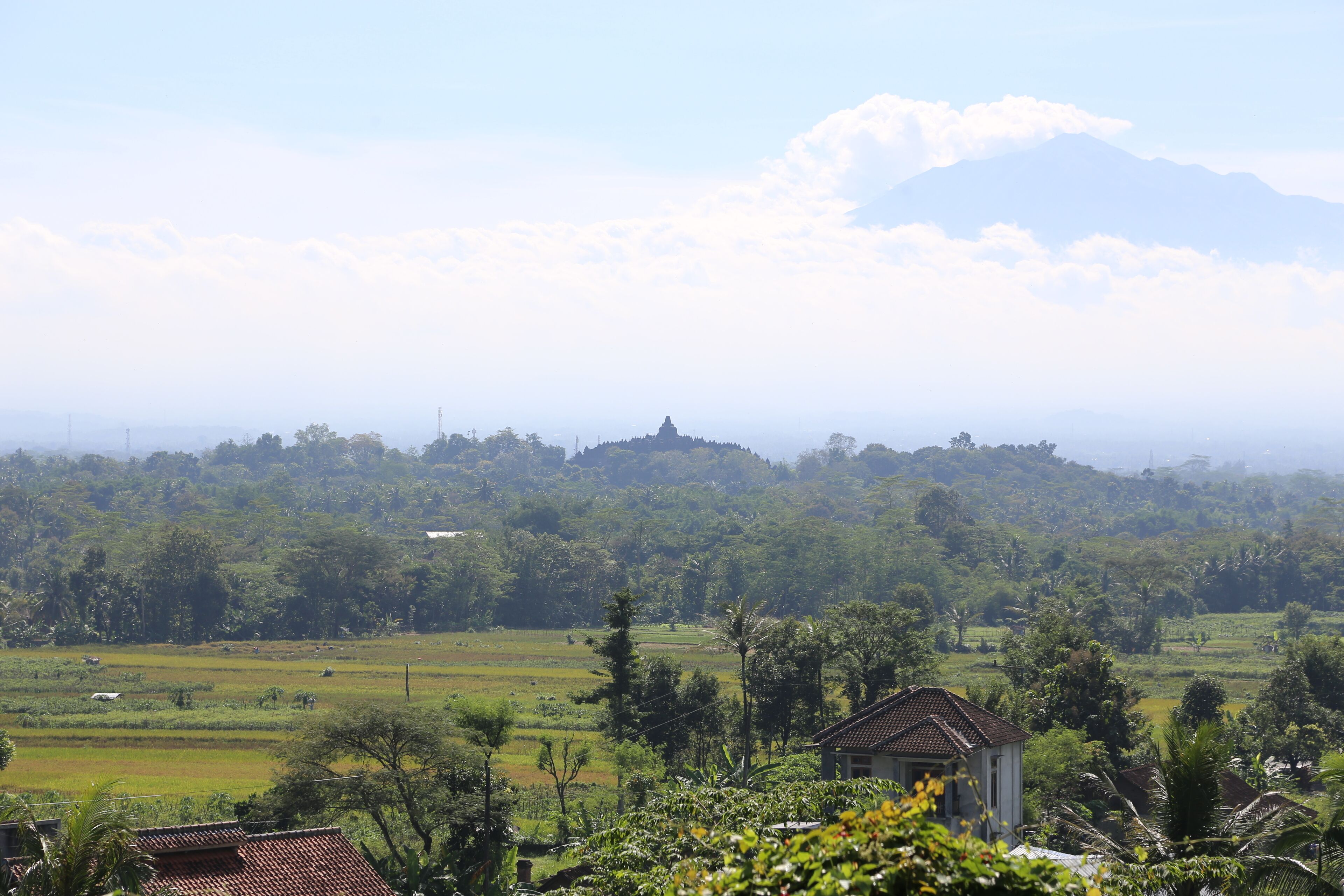 A rural landscape with a hilltop structure, lush greenery, and a small building with a tiled roof.