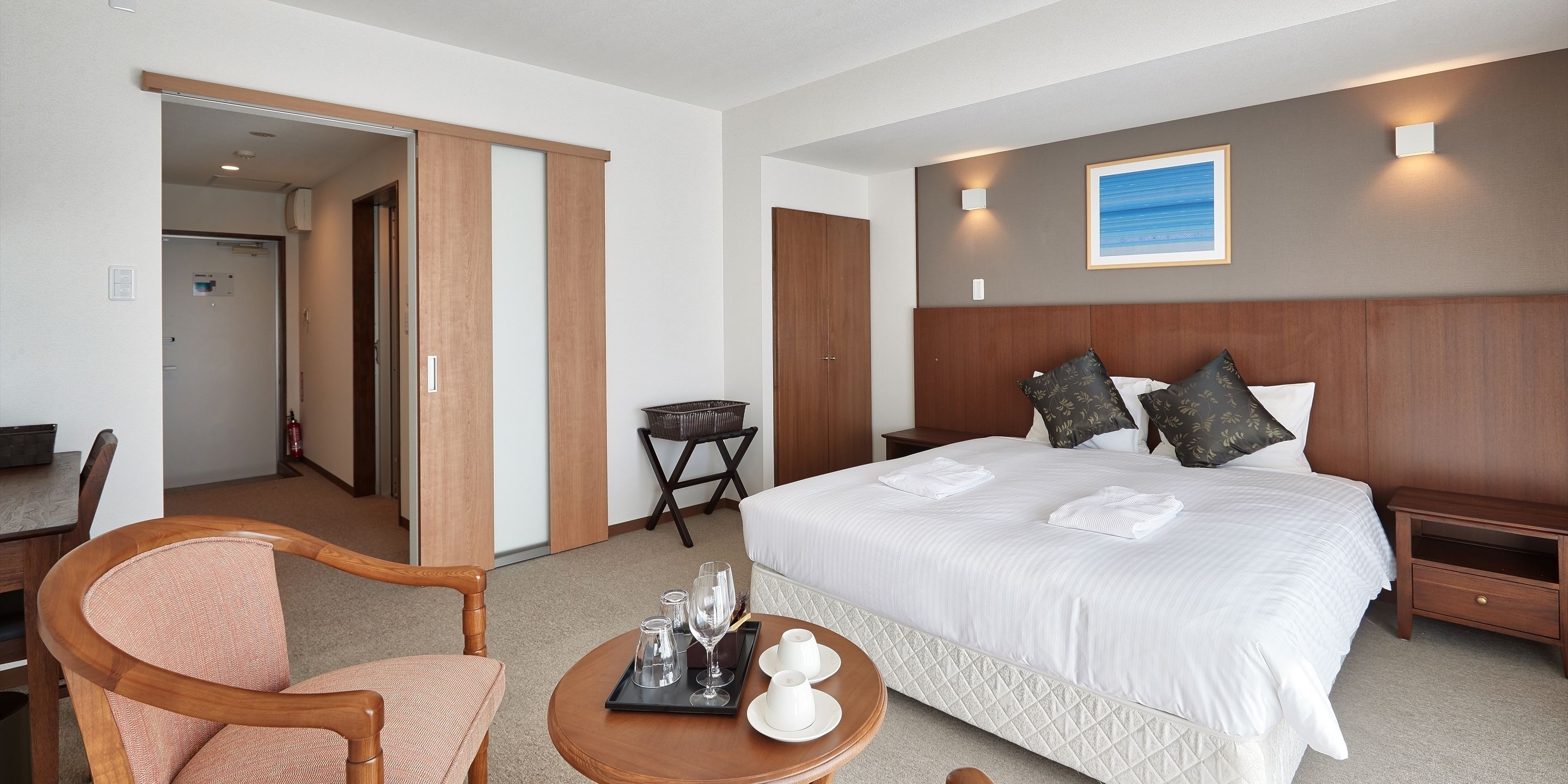 A hotel room with a large bed, a wooden chair, a small table with glasses and cups, and a painting on the wall.