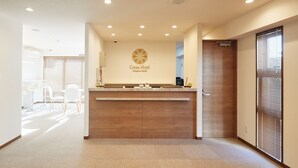 A hotel reception area with a wooden reception desk, a logo on the wall, and a view into a dining area with white chairs. - Creass Hotel TSUBOGAWA Marche (Naha)