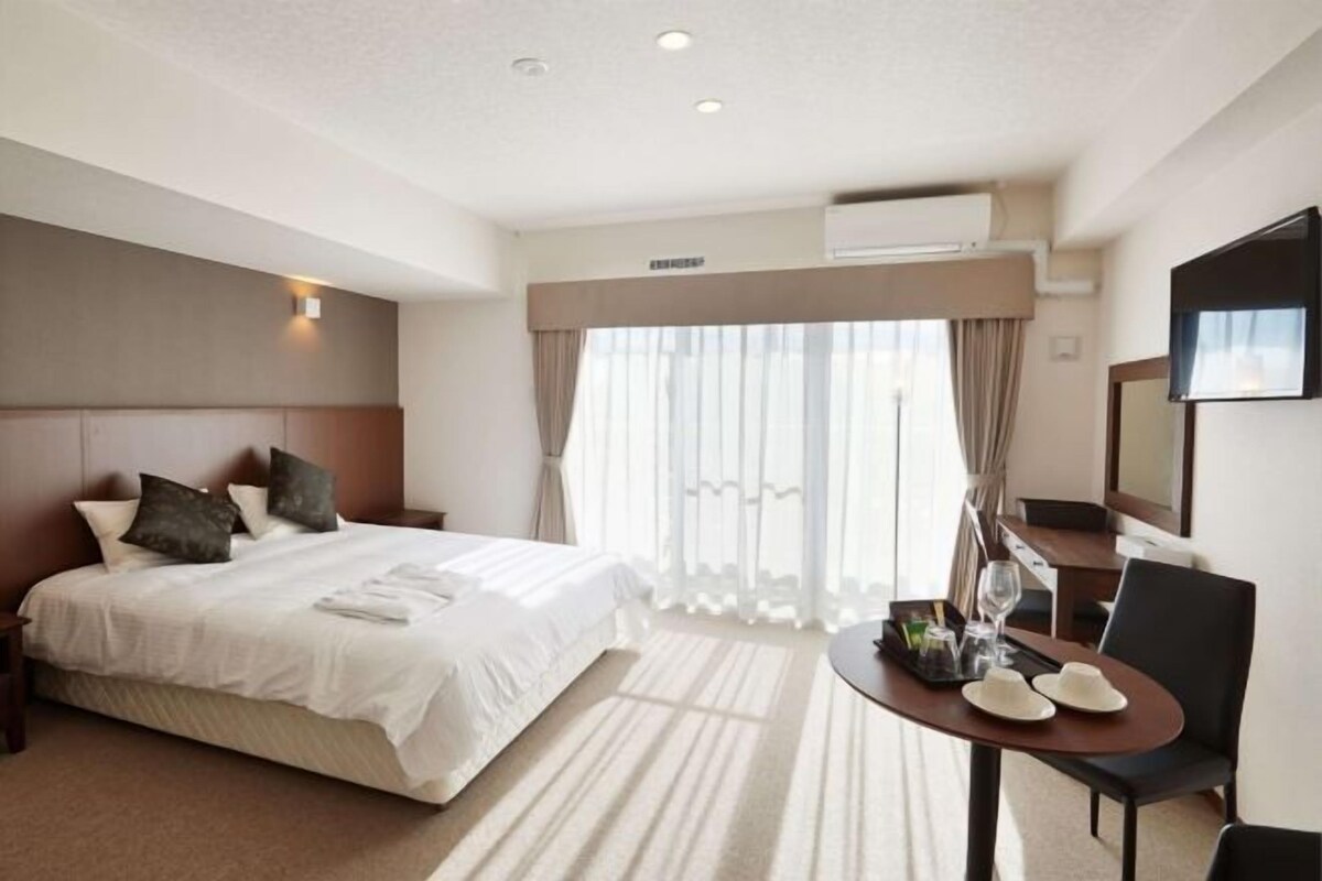 A hotel room with a large bed, a desk with a chair, and a window with curtains.