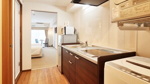 A compact kitchenette with a microwave, sink, and dishwasher. A bed and a small table with chairs are visible in the adjacent room. - Creass Hotel TSUBOGAWA Marche (Naha)