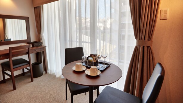 A hotel room with a round dining table set with a teapot, cups, and a tray. There is a wooden desk with a chair, a mirror, and a window with sheer curtains. - Creass Hotel TSUBOGAWA Marche (Naha)
