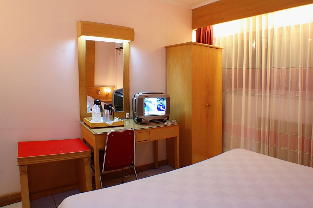 A hotel room with a bed, a desk with a television, a mirror, a red chair, and a wardrobe.