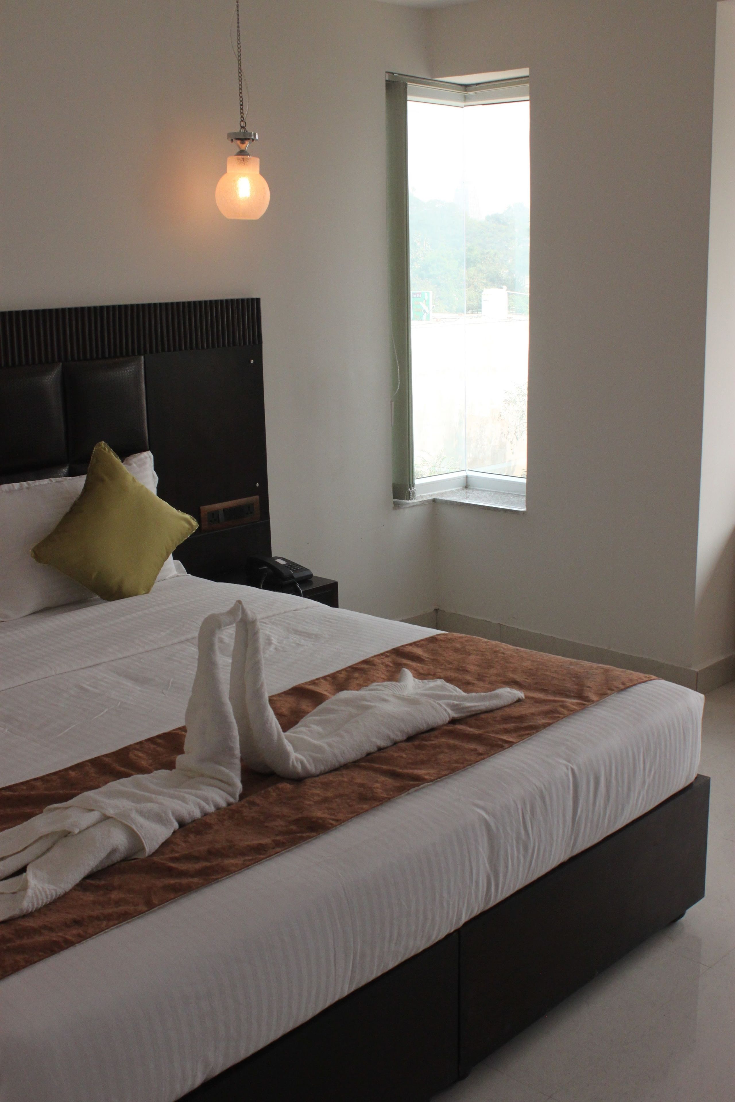 A neatly made bed with a brown accent, a bedside table with a phone, and a window with a view of greenery.