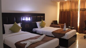 A hotel room with two beds, each with a headboard, pillows, and a blanket. There is a chair and a small table with a candle. - Maya Heritage (Gaya)
