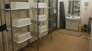 Una habitación con estanterías metálicas llenas de recipientes blancos, un lavabo con espejo y un pequeño cubo de basura.