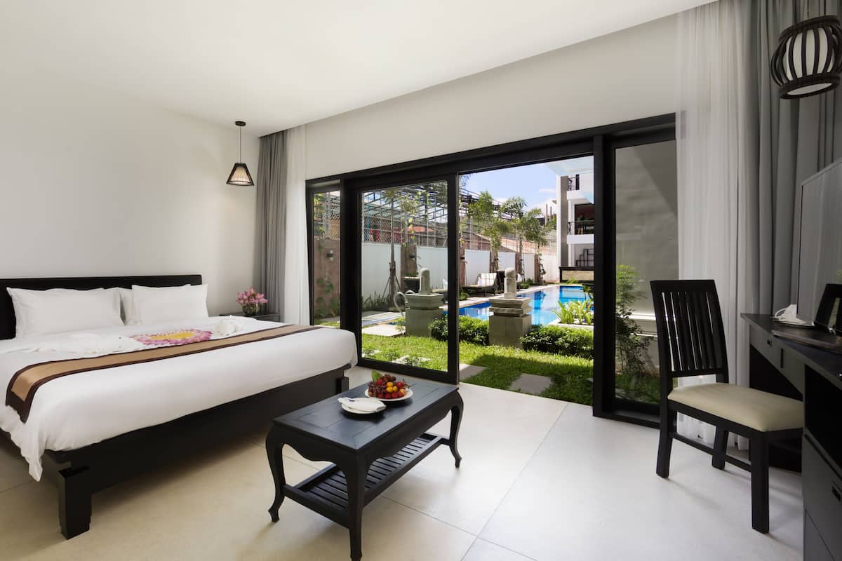 Ein modernes Hotelzimmer mit einem großen Bett, einem kleinen Tisch mit Früchten, einem Sessel und Blick auf einen Swimmingpool.