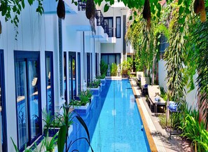 Un hotel moderno con una piscina larga, rodeado de abundante vegetación y un cielo azul claro.