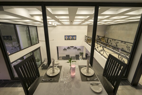 Un comedor con una mesa dispuesta para dos personas, un jarrón decorativo y vista al nivel inferior a través de un panel de vidrio.