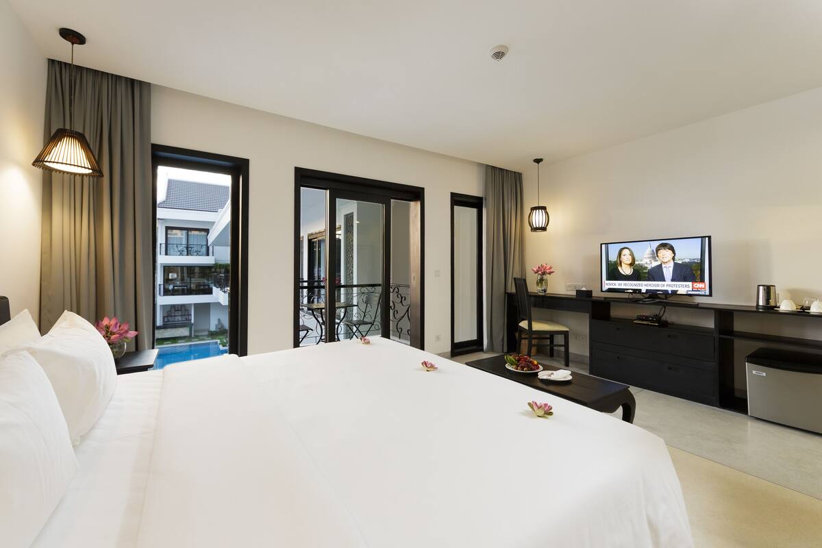 Ein Hotelzimmer mit einem großen Bett, einem Schreibtisch mit Computer, einem Fernseher und Blick auf ein Gebäude durch das Fenster.