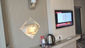 A hotel room with a TV mounted on the wall displaying a news channel, a bedside table with a kettle, a bottle of water, and a cup of coffee, and a mirror above the table. - Hotel Akdag (Usak)