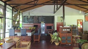 A spacious cafe with a high ceiling, wooden beams, and a menu board. There are wooden tables and chairs, and a counter with a beverage dispenser. - Highland Lodge Hotel (Yangon)