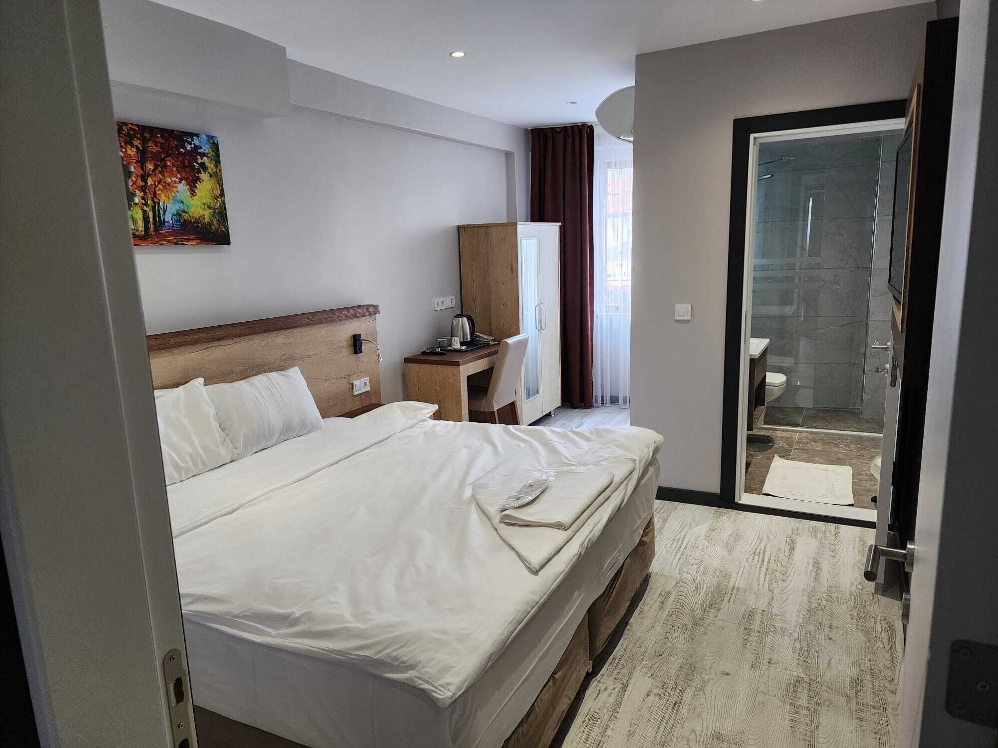 A hotel room with a large bed, a desk with a chair, a wardrobe, and a glass door leading to a bathroom.