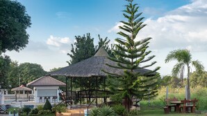 Ein Pavillon mit strohgedecktem Dach, umgeben von üppiger Vegetation, einem Pool und einem kleinen Gebäude mit Ziegeldach.