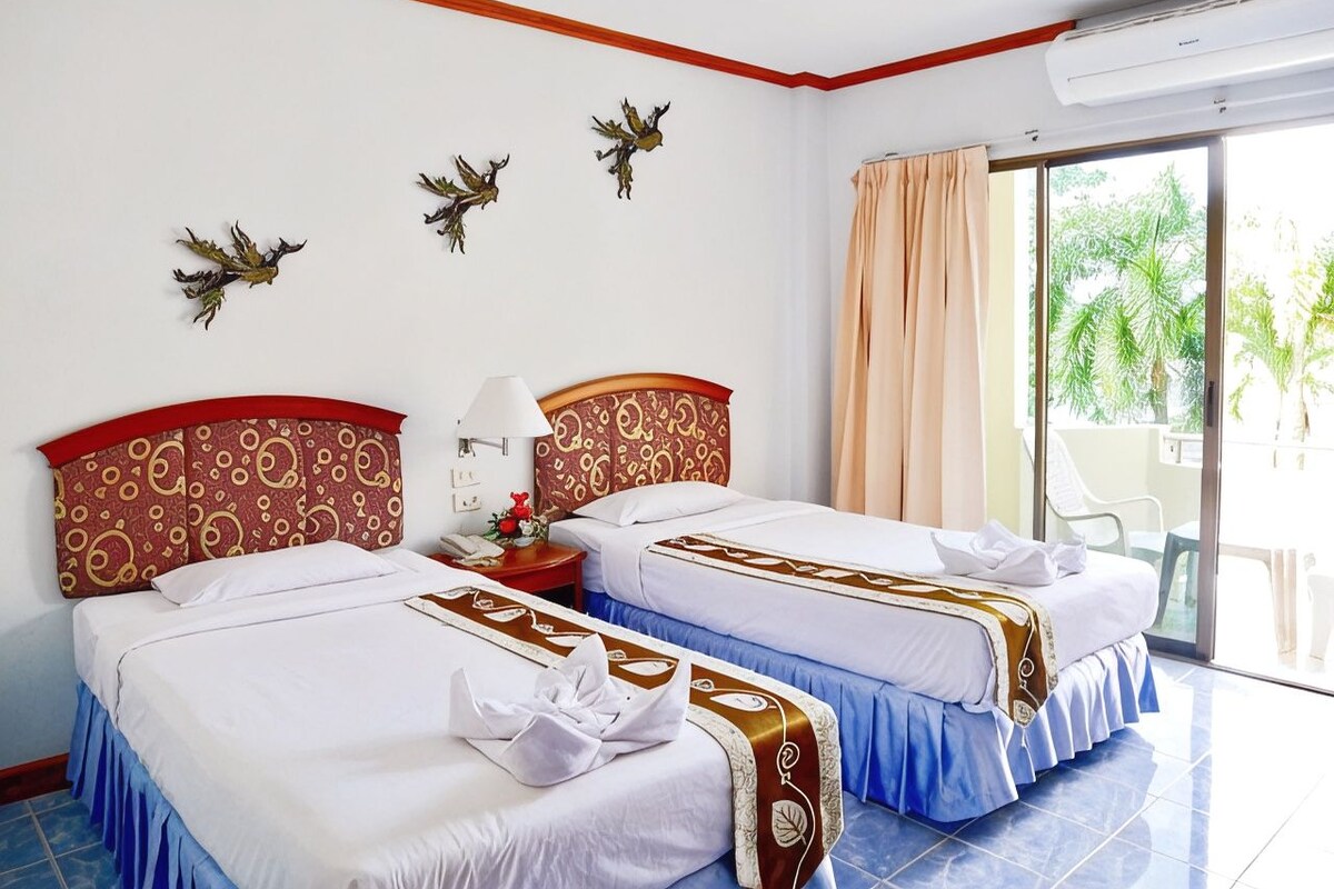 Une chambre d’hôtel avec deux lits, une tête de lit rouge et dorée, une grande fenêtre donnant sur des palmiers et un appareil de climatisation.