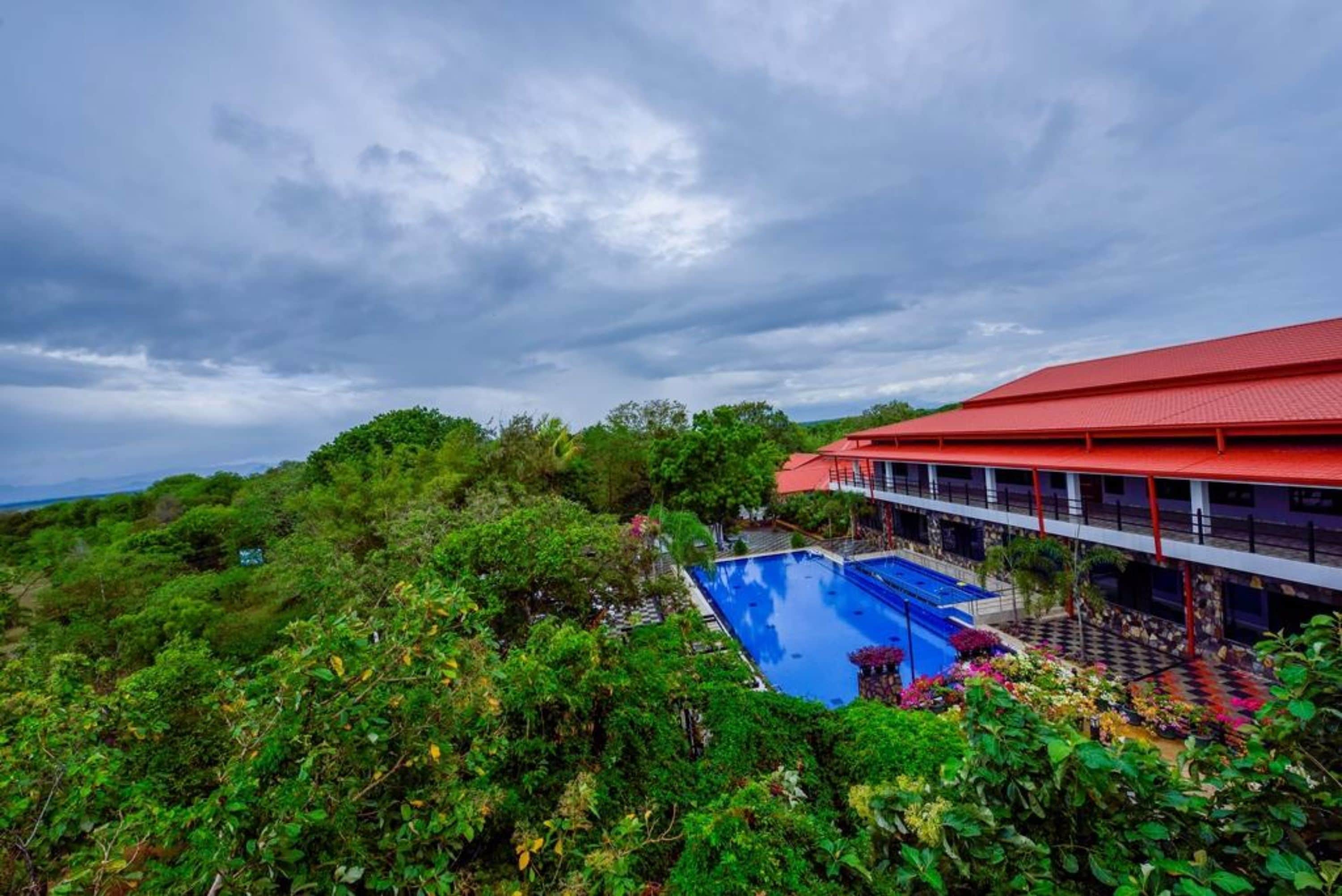 A hotel with a swimming pool surrounded by lush greenery and a red-roofed building.