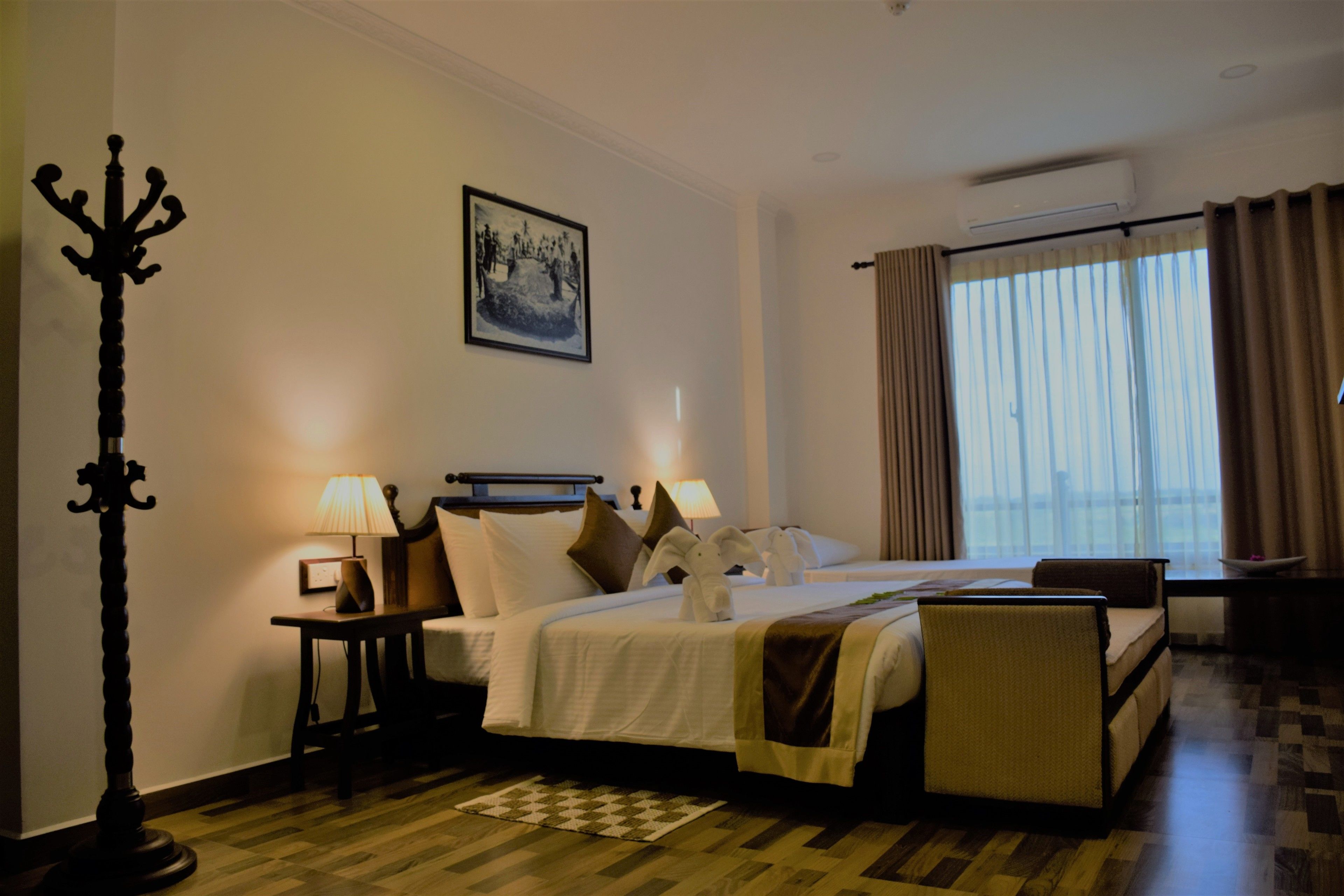 Ein Hotelzimmer mit einem Bett, zwei Nachttischlampen, einem Nachttisch, einem Sessel, einer Stehlampe, einem Fenster mit Vorhängen und einem gerahmten Bild an der Wand.