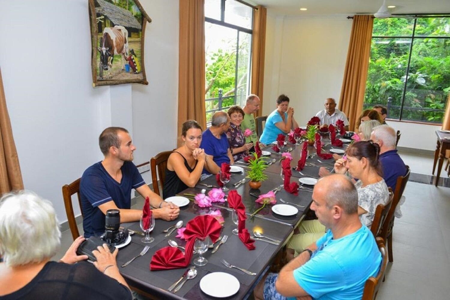 Un groupe de personnes est assis autour d’une table dans une pièce bien éclairée, dotée de grandes fenêtres, avec un tableau encadré au mur et une table dressée avec des serviettes rouges et de la verrerie.