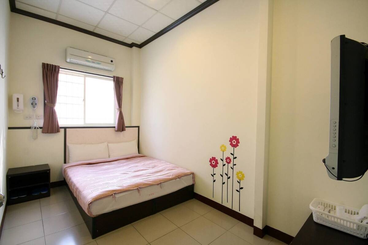 A bedroom with a bed, an air conditioner, a telephone, a window with curtains, and a wall-mounted TV.
