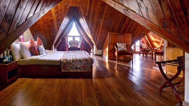 Une chambre mansardée chaleureuse, avec un plafond en pente en bois, un lit recouvert d’une literie à motifs floraux et un petit coin salon avec des chaises et une table.