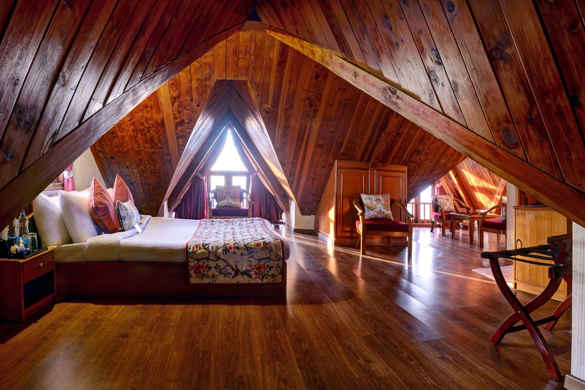 A cozy attic bedroom with a wooden sloped ceiling, a bed with floral bedding, and a small seating area with chairs and a table.