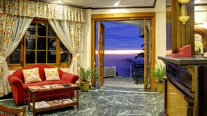 Un salon chaleureux avec un canapé rouge, une table basse et une vue sur les montagnes au coucher du soleil, visible à travers une porte ouverte.