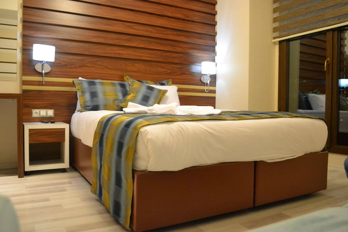 Quarto de hotel moderno com cabeceira de madeira, cama com manta listrada, duas luminárias de cabeceira e uma mesinha de apoio.