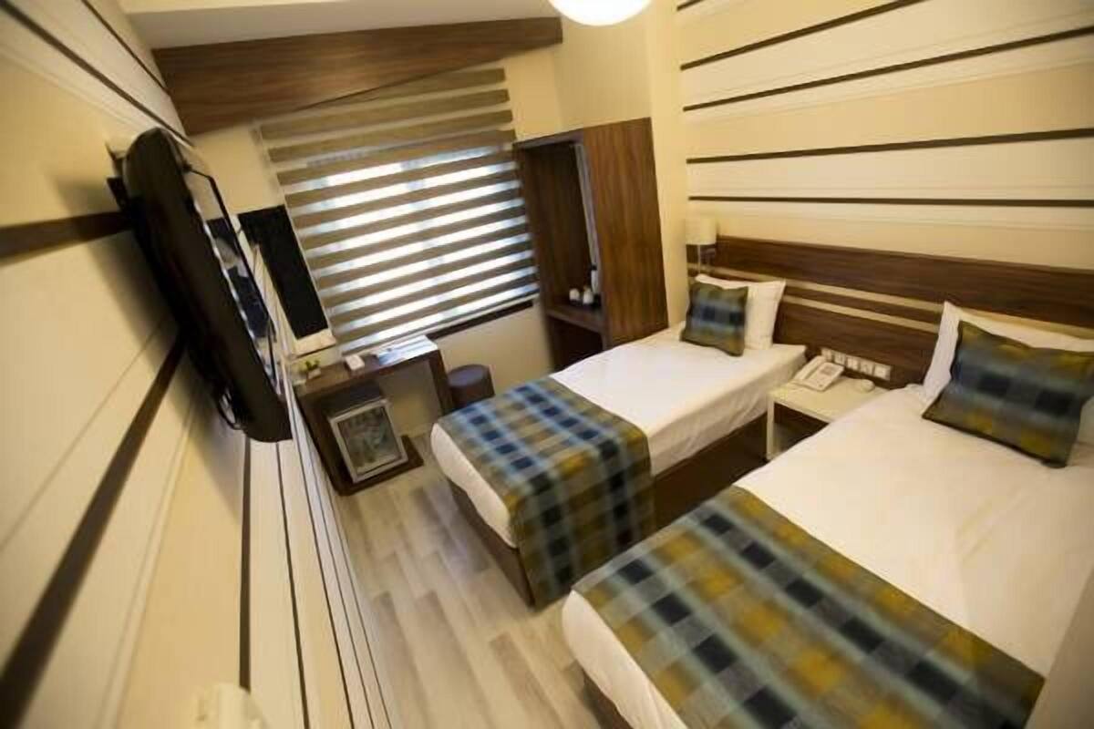 Quarto de hotel com duas camas, televisão, mesa de cabeceira e janela com persianas.