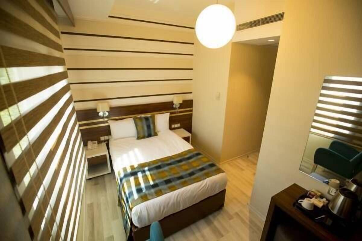 Quarto de hotel moderno com parede listrada, cama com colcha xadrez, mesa de cabeceira com abajur e pequena escrivaninha com cafeteira.