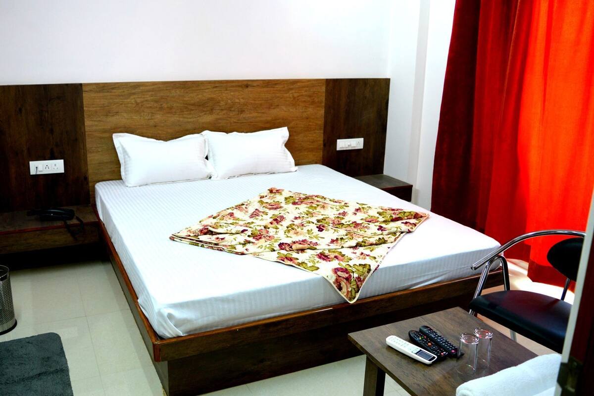 Habitación de hotel con cama de madera, ropa de cama blanca, un plaid floral, una mesita de noche con control remoto y una silla.