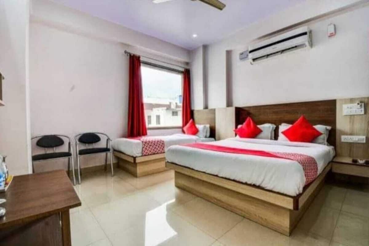 Habitación de hotel con dos camas individuales, una mesita pequeña, dos sillas, un ventanal con cortinas rojas y un equipo de aire acondicionado.