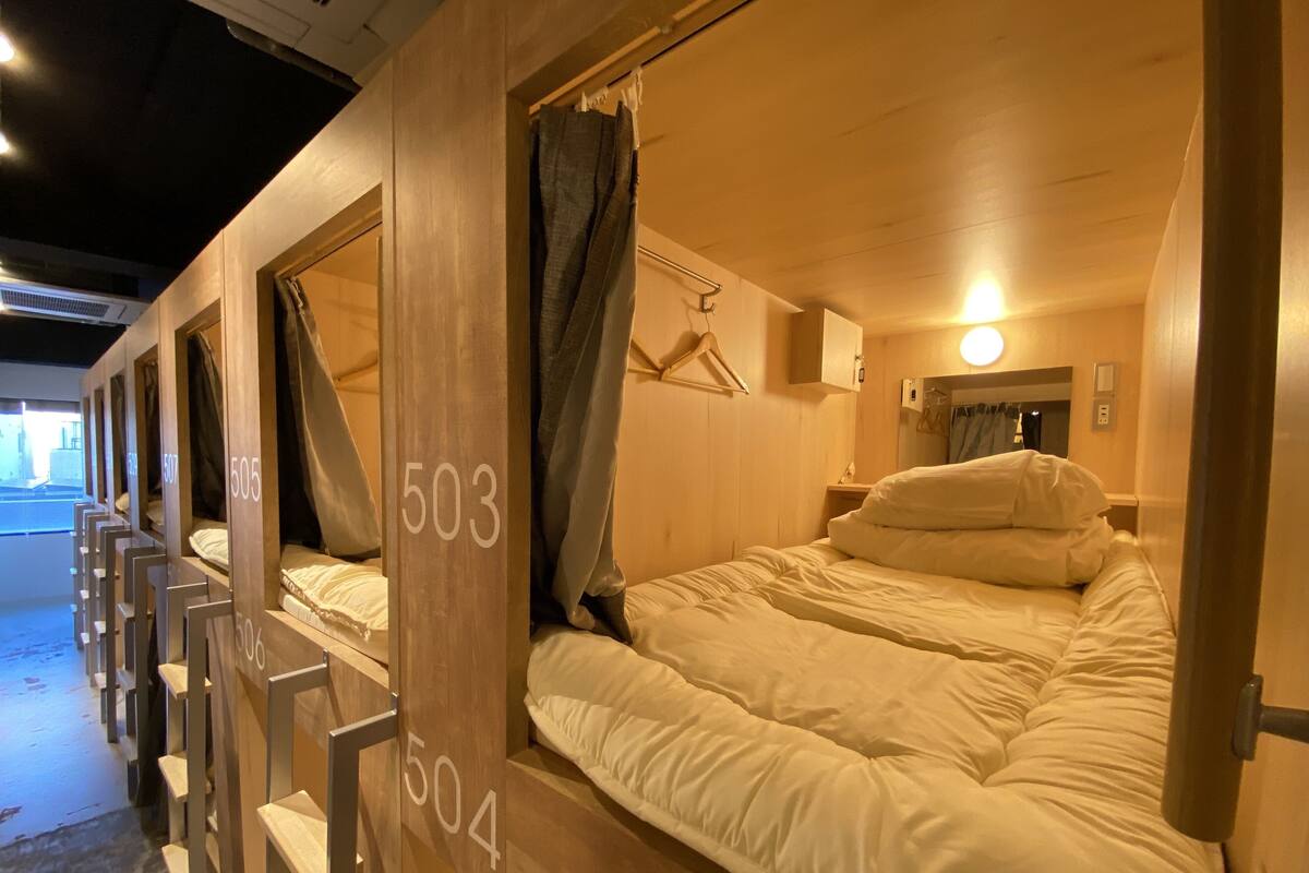 Habitación de hotel con literas, puertas numeradas, cama con ropa blanca, mesita de noche y espejo montado en la pared.