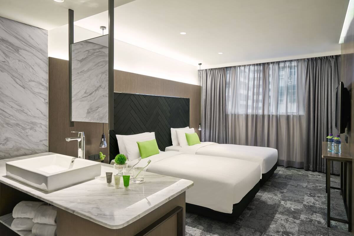 Une chambre d’hôtel moderne avec une grande salle de bain en marbre, deux lits avec une literie blanche et des oreillers verts, un grand miroir et un téléviseur à écran plat posé sur un meuble en bois.