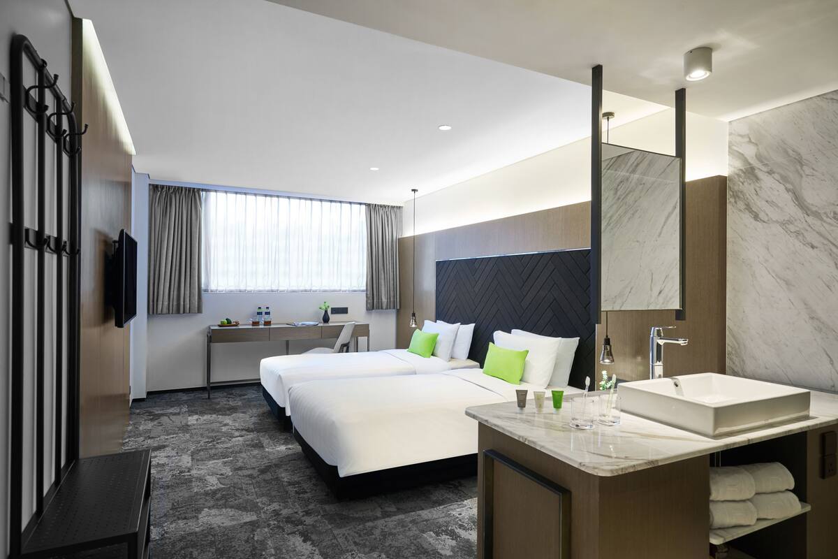 Une chambre d’hôtel moderne avec deux lits, un grand miroir, une salle de bain avec un lavabo et un bureau avec une chaise.