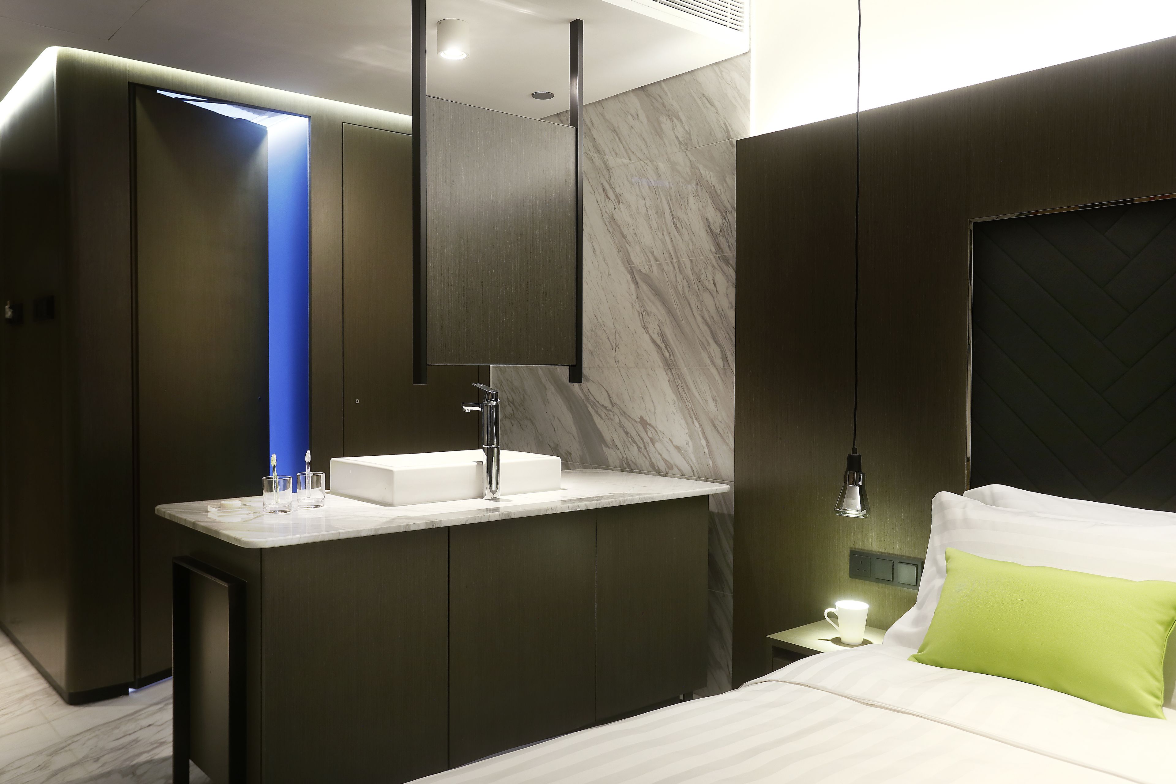 A modern hotel room with a built-in vanity, a bed with a green pillow, and a nightstand with a lamp.