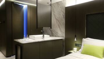 A modern hotel room with a built-in vanity, a bed with a green pillow, and a nightstand with a lamp.