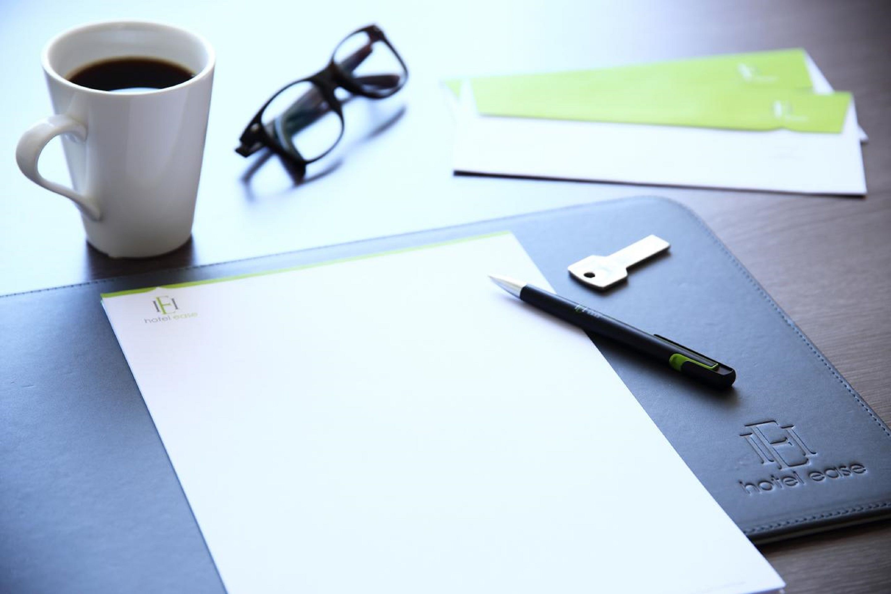 A desk with a white mug, black glasses, a pen, a key, and a notebook with a hotel logo.