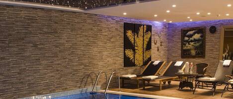 Una piscina interior con sillones de descanso, una mesa con una bebida y una botella, y detalles decorativos en la pared.