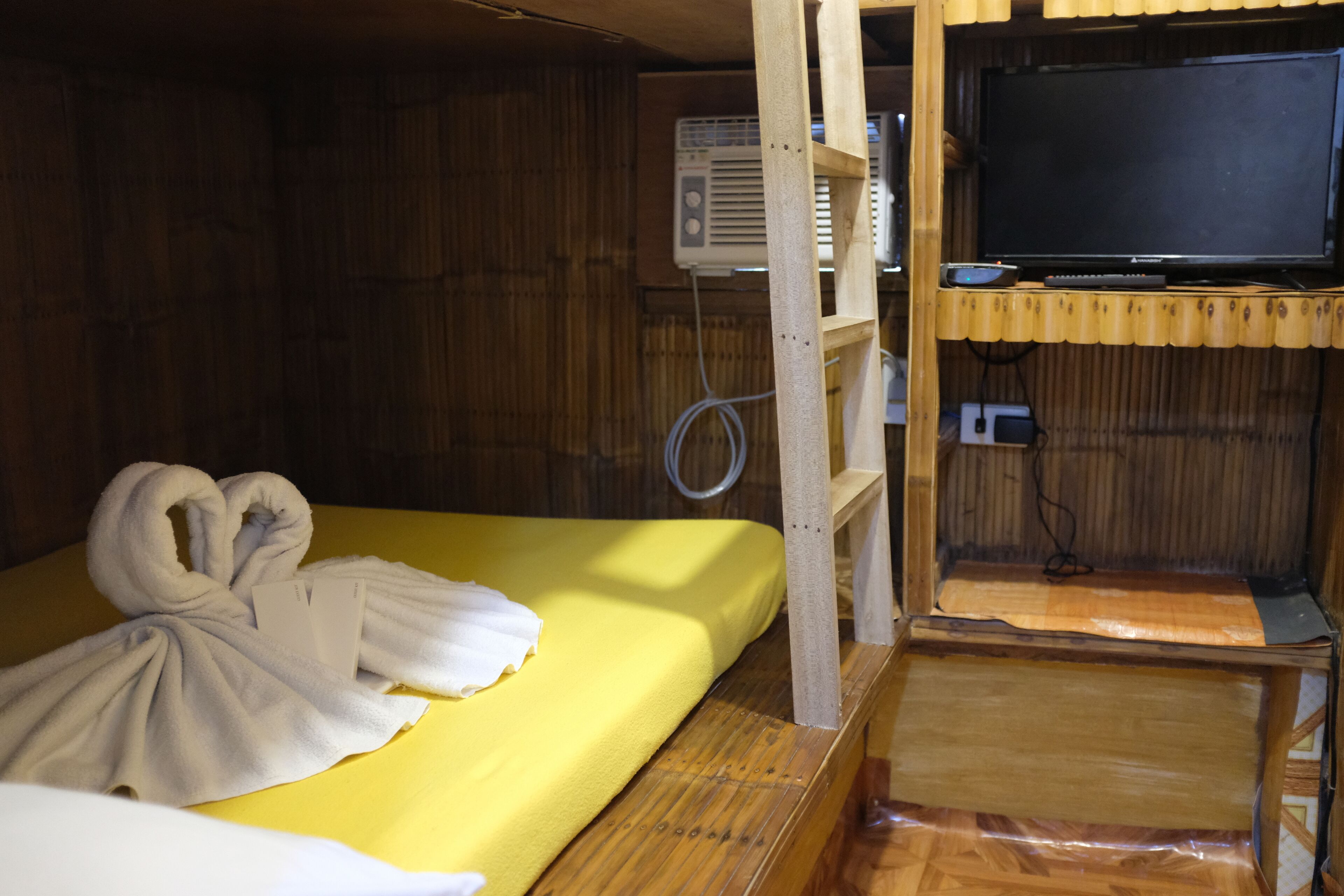 A compact room with a bed, a television on a wooden stand, and an air conditioning unit.
