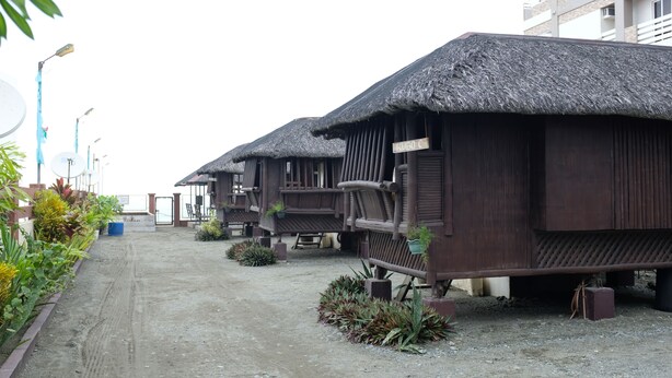 A row of thatched-roof huts with wooden railings, arranged along a dirt path with potted plants and a building in the background. - Nipa Hut Villa by AMCO Extension (Baler)