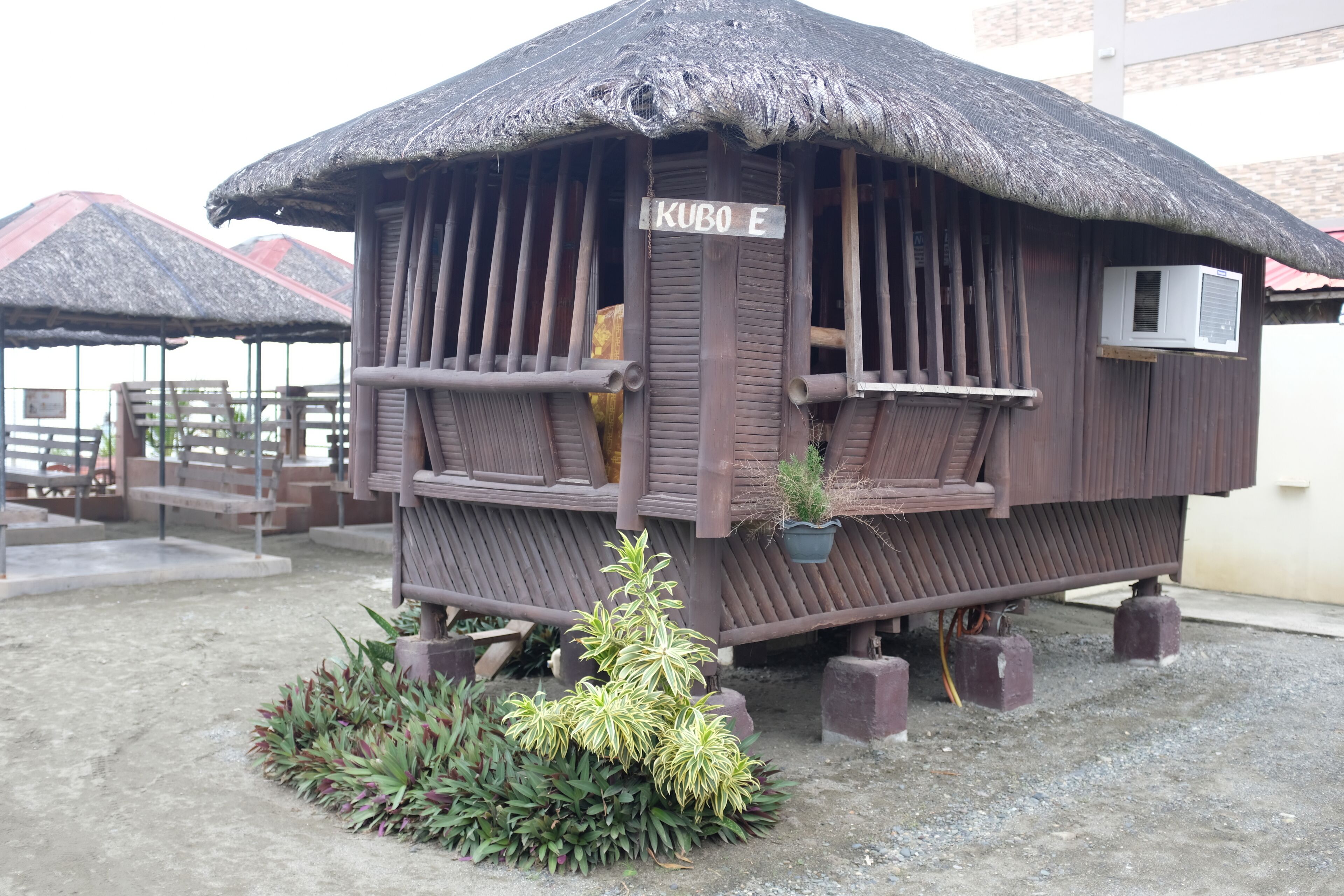 A small wooden structure with a thatched roof labeled "KUBO E," featuring a small balcony and a potted plant.