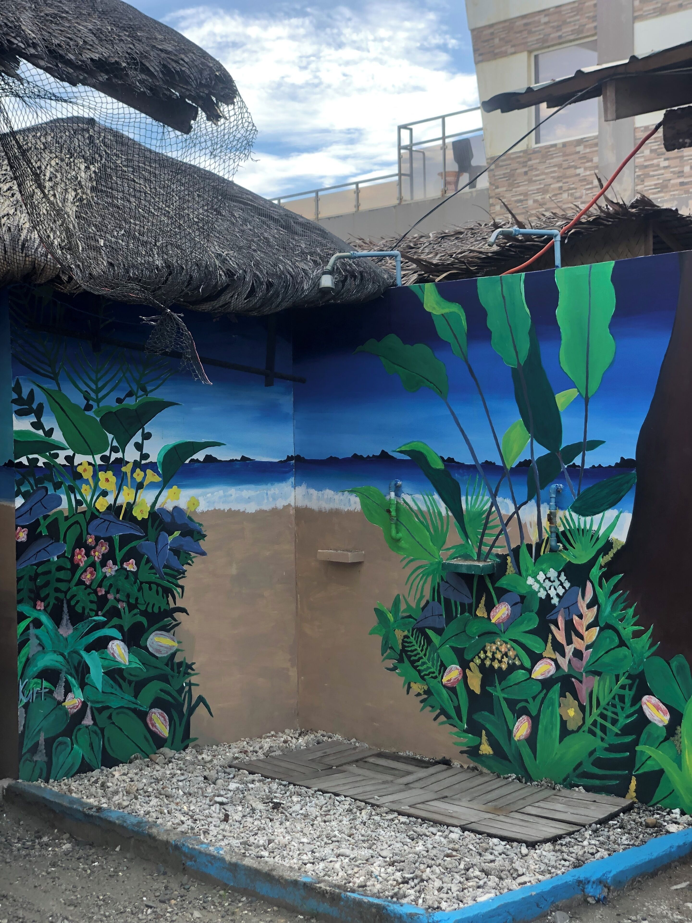 Un mural que representa una escena tropical con plantas y flores, una estructura de techo de paja y un cielo azul.