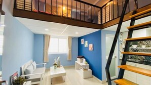 A modern living room with a blue accent wall, a white sofa, a dining table, and a staircase with a wooden railing. - The Homee Cozy Modern Studio Apartment (Ho Chi Minh City)