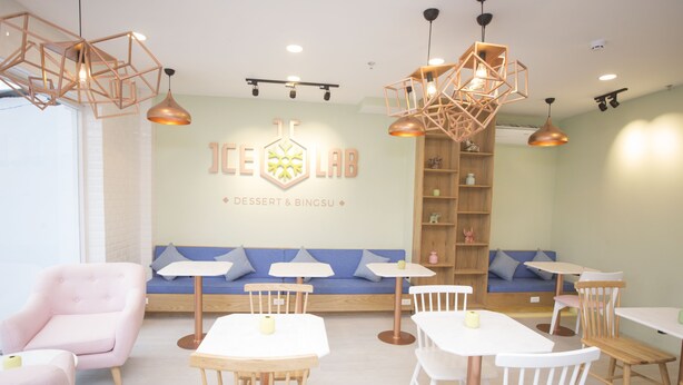 A modern cafe interior with a central seating area, white tables, and chairs, and a wall with the text "ICE LAB DESSERT & BINGSU". - The Homee Cozy Modern Studio Apartment (Ho Chi Minh City)