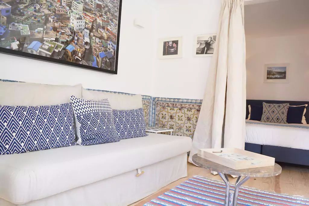 A modern living room with a white sofa, patterned cushions, a glass coffee table, and a large cityscape painting on the wall.