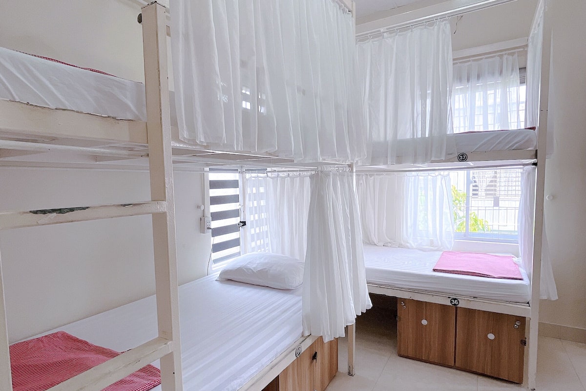 A dormitory room with bunk beds, white curtains, and a window with blinds.