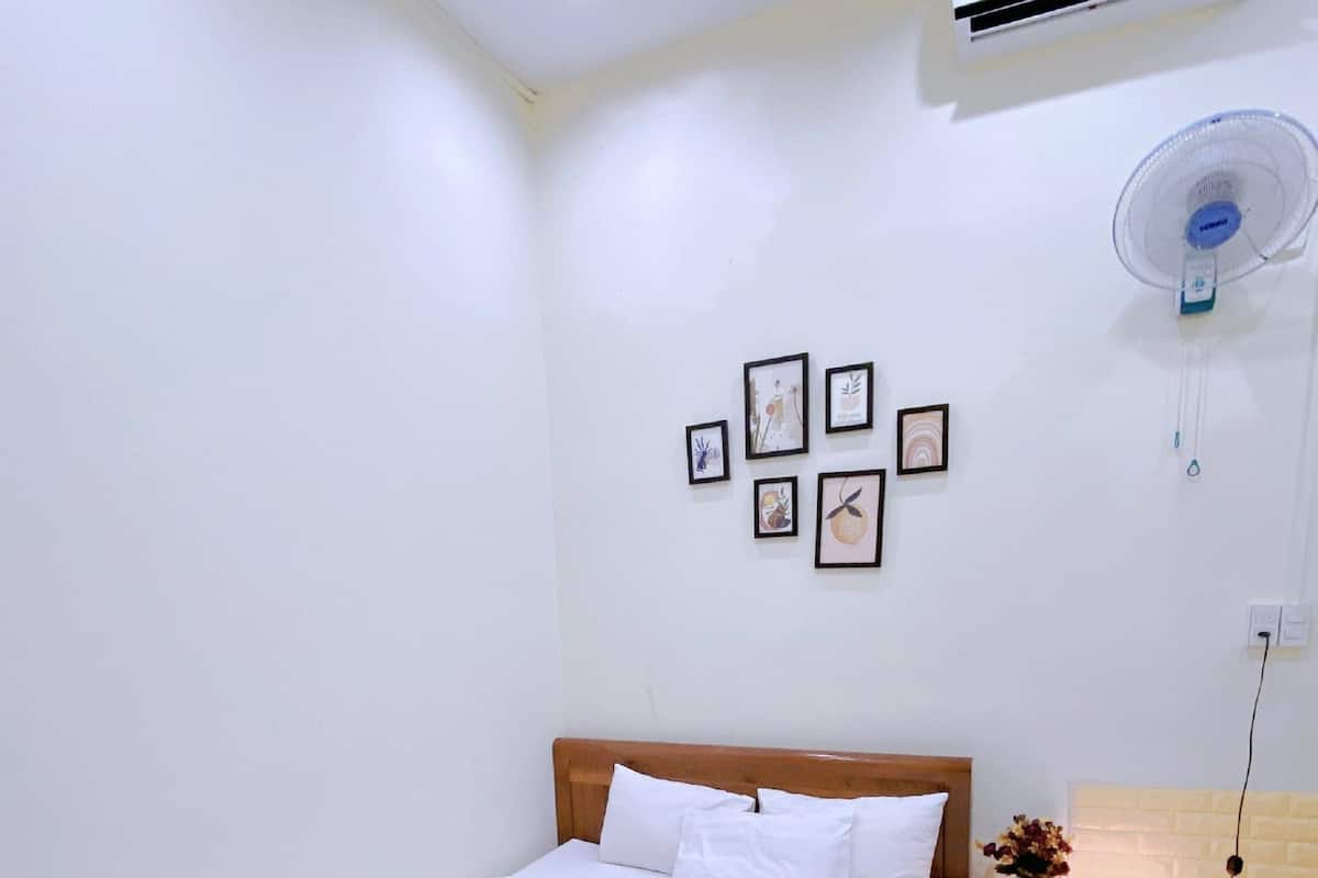 A neatly made bed with white linens and towels, a wooden headboard, a bedside table with a lamp and a vase of flowers, and a wall-mounted air conditioner and fan.