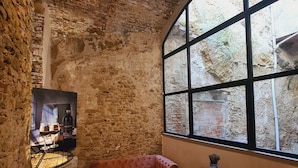 Una habitación rústica con paredes de ladrillo visto, un amplio ventanal de arco, un sofá rosa, un banco de madera y una pequeña mesa con una planta.