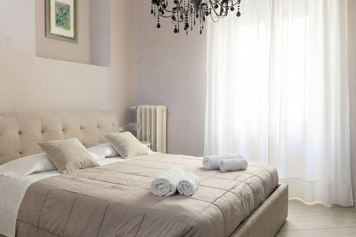 A bedroom with a large bed, a bedside table, a radiator, and a chandelier.